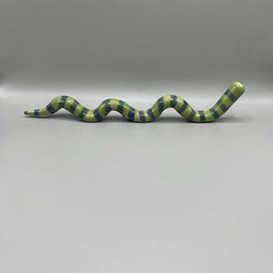 May include: A ceramic snake figurine with a green, blue, and cream striped pattern. The snake is in a curved pose, with a rounded head and a tapered tail. The figurine is on a plain, light gray background.