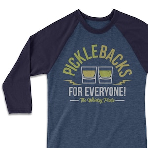 May include: A blue and navy blue baseball tee shirt with the text "Picklebacks for Everyone! The Whiskey Pickle" in yellow and white lettering. Two yellow and green shot glasses are pictured above the text.