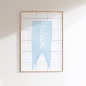 Puede incluir: Un estampado enmarcado con una pancarta azul claro con la palabra "Kentucky" en escritura blanca. La pancarta está sobre un fondo a cuadros azul claro y blanco. El marco es de color marrón claro.
