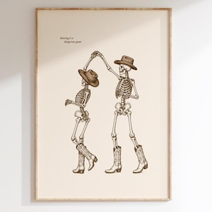 Puede incluir: Una lámina de arte enmarcada sobre un fondo crema con un dibujo lineal en marrón oscuro de dos esqueletos bailando. Ambos esqueletos llevan sombreros de vaquero y botas de estilo western. El texto "dancing is a dangerous game" aparece impreso en la parte superior izquierda.