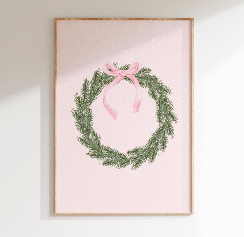 May include: A framed print featuring a watercolor illustration of a Christmas wreath. The wreath is made of green pine needles and adorned with a pink ribbon. The background is a soft pink with subtle white speckles, creating a festive and elegant aesthetic.