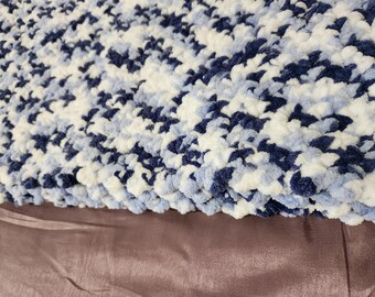 May include: A close-up of a soft, textured blanket in shades of navy blue, light blue, and white. The blanket has a plush, puffy appearance, and is likely made of a cozy material. The blanket is resting on a brown surface.