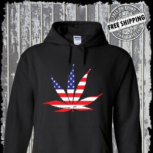 Op de afbeelding: Zwart hoodie met een marihuanablad ontwerp met de kleuren van de Amerikaanse vlag. De tekst "Kinsman Trading Co. LLC" staat onderaan de afbeelding.