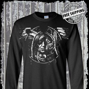 Może przedstawiać: Czarny T-shirt z długim rękawem z nadrukiem graficznym przedstawiającym Indianina z twarzą wilka, otoczonego piórami i łapaczem snów. Grafika jest w odcieniach szarości.