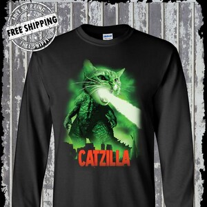 May include: Black long-sleeved t-shirt featuring a green and white graphic of a cat with a Godzilla-like appearance, breathing fire. The graphic text reads "CATZILLA".