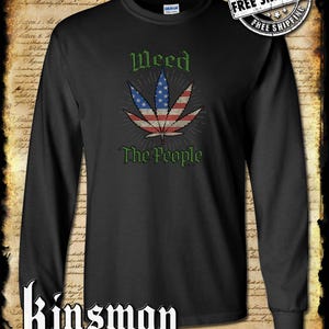 Op de afbeelding: Zwart langarm T-shirt met een groen marihuanablad ontwerp. Het blad is een Amerikaanse vlag met rode en witte strepen en witte sterren. De tekst "Weed The People" is onder het blad gedrukt.