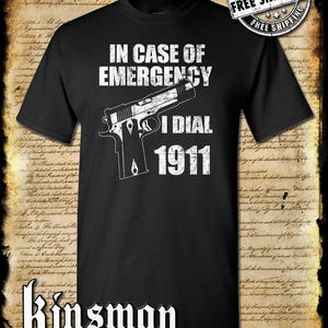 Pode incluir: Camiseta preta com um gráfico branco de uma arma de fogo e o texto "IN CASE OF EMERGENCY I DIAL 1911". O texto está em uma fonte desgastada. A camiseta tem um logotipo branco "Kinsman Trading Co. LLC" na parte inferior.