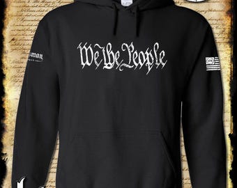 May include: Black hooded sweatshirt with the phrase "We the People" in white gothic lettering across the chest. The sleeve features a small American flag graphic. The brand name "Kinsman trading co llc" is printed at the bottom.