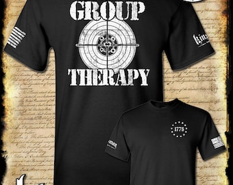 Puede incluir: Camiseta negra con texto blanco que dice "This is what I call group therapy" y un objetivo con agujeros de bala. La parte posterior de la camiseta tiene un círculo blanco con el texto "1776" y estrellas alrededor.