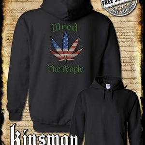 Op de afbeelding: Zwarte hoodie met een cannabisbladontwerp in de kleuren van de Amerikaanse vlag. De tekst "Weed The People" staat boven en onder het blad. De merknaam "Kinsman trading co llc" staat onderaan.
