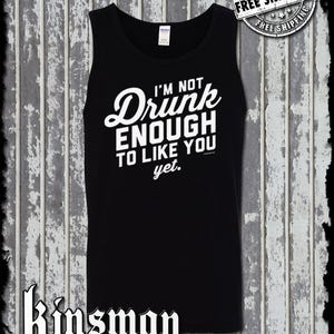 May include: Black tank top with the text "I'M NOT Drunk ENOUGH TO LIKE YOU yet." in white. The tank top has a crew neck and a sleeveless design. The Kinsman trading co. logo is at the bottom.