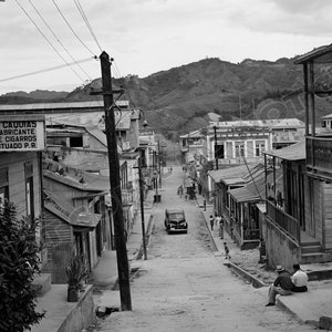 May include: A black and white photo of a street in a small town. The street is lined with wooden buildings, some with balconies. A car is driving up the street. The sign on the building reads "R. Caudias Fabricante de Cigarros Utuado P.R."