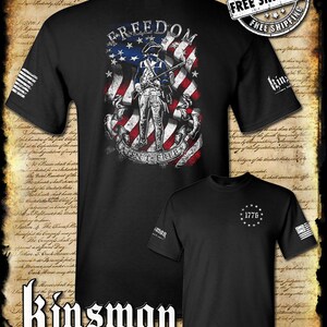May include: Black t-shirts with patriotic designs. One shirt features a Revolutionary War soldier in front of an American flag with the word "FREEDOM". The other shirt has "1776" surrounded by stars. The brand name "Kinsman" is also visible.