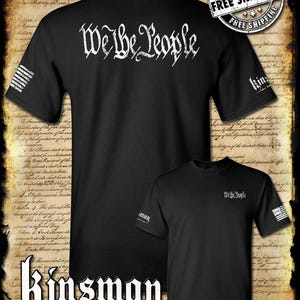 May include: Black t-shirt with a white "We the People" graphic on the back and a small American flag on the left sleeve. The front of the shirt has a small "We the People" graphic on the right side and a small American flag on the right sleeve. The shirt is on a background of a vintage document.