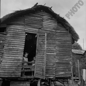 May include: Two children stand in the doorway of a wooden shack with a thatched roof. The shack is raised off the ground on wooden posts. The children are looking out at the viewer.