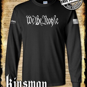 Puede incluir: Camiseta negra de manga larga con una impresión blanca de "We the People" en una fuente de estilo vintage. La camiseta tiene un pequeño parche de bandera estadounidense en la manga derecha. El texto "Kinsman Trading Co. LLC" está impreso debajo de la imagen.