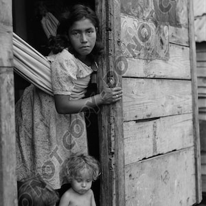 May include: A black and white photograph of three children peeking out from behind a wooden door. The oldest child, a girl, is holding the door open with her hand. The two younger children are sitting on the floor.