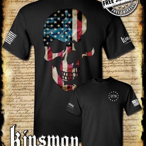 May include: Black t-shirts featuring a skull design with an American flag motif. One shirt displays a skull with the flag's stars and stripes, while the other has "1776" and a star pattern. The brand name "Kinsman" is also visible.