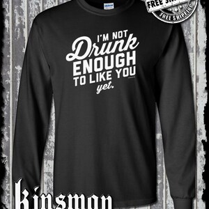 May include: Black long-sleeve shirt with the text "I'M NOT Drunk ENOUGH TO LIKE YOU yet." in white script. The shirt also features the "GILDAN" brand label and the "Kinsman trading co llc" logo.