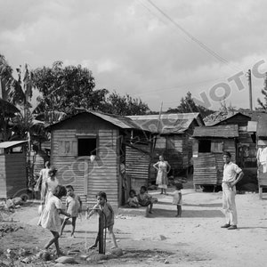 May include: A black and white photo of a group of children playing in front of a row of small wooden houses. The houses are simple and have corrugated metal roofs. There are banana trees in the background. The children are dressed in simple clothing and are playing with a water pump.