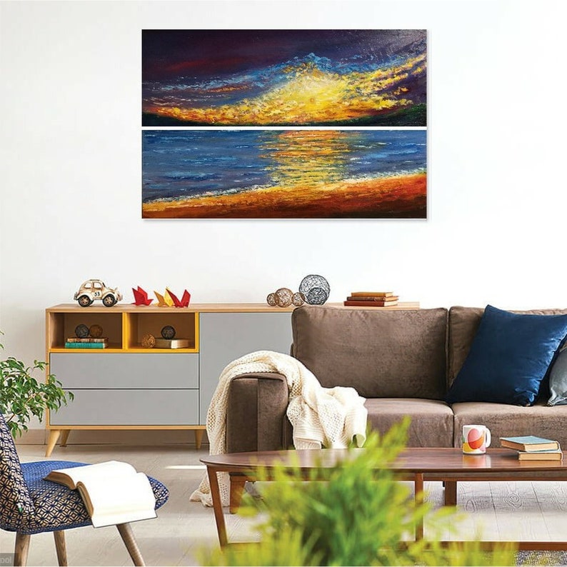 Pode incluir: Uma pintura abstrata de duas pe&ccedil;as que retrata um p&ocirc;r do sol sobre um corpo de &aacute;gua. A pe&ccedil;a superior mostra um c&eacute;u vibrante amarelo e laranja com nuvens azul escuro. A pe&ccedil;a inferior mostra um oceano azul com um reflexo dourado do p&ocirc;r do sol. A pintura &eacute; emoldurada e pendurada em uma parede branca.