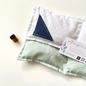 May include: A green and white rice pack with a blue denim triangle patch. The pack is labeled "soothing packs" and includes information about aromatherapy rice packs, essential oil roller blends, aromatherapy diffusers, and jewelry.