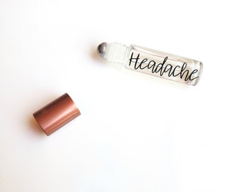 May include: A clear glass roller bottle with a silver roller ball and a black label that says "Headache". The bottle is sitting on a white surface next to a copper-colored cap.