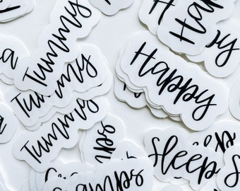 Puede incluir: Una colección de pegatinas blancas con texto escrito a mano en negro. El texto incluye "Happy", "Tummy", "Cramps" y "Sleep".