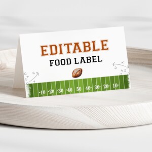 May include: A white, folded card with the words "EDITABLE FOOD LABEL" in brown and black. A football graphic sits above a green field with yard lines. The card is displayed on a light-coloured wooden tray.