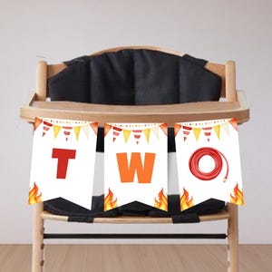 May include: A high chair decorated with a banner that says "TWO" with a fire truck theme. The banner has red, orange, and yellow accents, including flames and a fire hose graphic. The high chair is made of wood and has a black seat.