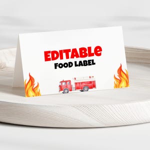 May include: A white, folded card with the words "EDITABLE FOOD LABEL" in red and black. A red fire truck illustration is featured below the text. Flames are on either side of the card. The card is on a light-colored wooden tray.