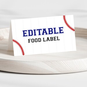 May include: A white baseball-themed food label with the word "EDITABLE" in blue and the words "FOOD LABEL" in black. Red baseball stitching accents the corners. The label is displayed on a light-colored wooden tray.