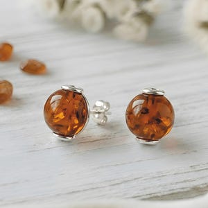 Può includere: Un paio di orecchini a lobo in ambra con finiture argentate. Gli orecchini presentano una pietra d'ambra rotonda e lucida. Lo sfondo include piccole pietre d'ambra e fiori bianchi, suggerendo un tema di gioielli.