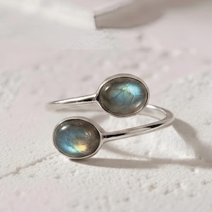 Op de afbeelding: Een zilveren ring met twee ovale labradorietstenen. De ringband loopt rond de vinger, met een steen aan de bovenkant en de andere aan de onderkant. De stenen hebben een blauwgroene iriserende glans.