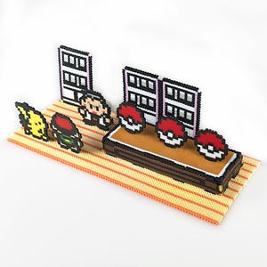 May include: A pixel art diorama depicting scenes from the Pokémon franchise. The scene includes a yellow Pikachu, a trainer, and several Poké Balls on a brown platform, all set against a backdrop of pixelated buildings.