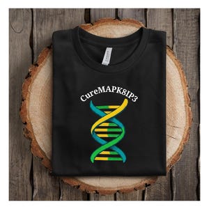 May include: Black t-shirt featuring the text "CureMAPK8IP3" above a DNA helix design in teal, yellow, and green. The shirt is folded and placed on a wooden surface.
