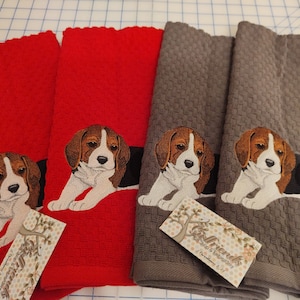 May include: Set of three kitchen towels featuring embroidered beagle dog designs. Two towels are red and one is gray. The towels have a waffle weave texture. The towels have a tag that says "Gifted Friends" with a floral design.