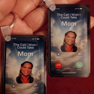 May include: Two smartphone-shaped keyrings, each featuring a photo and the text "The Call I Wish I Could Take Mom." The screens show a sky background and call buttons. The keyrings are held by a hand.