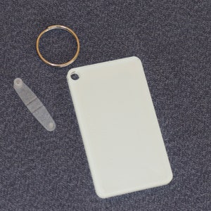 May include: A white rectangular tag with a rounded corner and a hole in the top corner, a gold-colored key ring, and a small, clear plastic piece with two holes and a rectangular indentation. The items are on a dark gray textured surface.