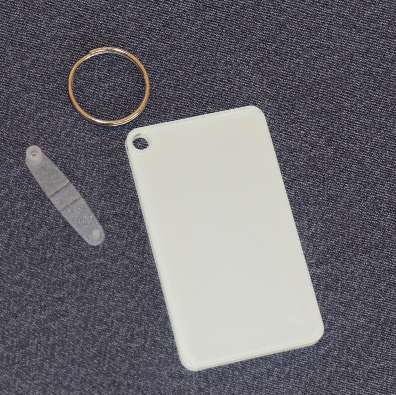 May include: A white rectangular tag with a rounded corner and a hole in the top corner, a gold-colored key ring, and a small, clear plastic piece with two holes and a rectangular indentation. The items are on a dark gray textured surface.