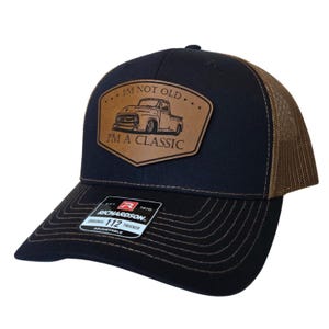 May include: Black and brown trucker hat with a brown leather patch featuring a classic truck illustration and the text "I'M NOT OLD I'M A CLASSIC." The hat has a curved brim and adjustable closure.