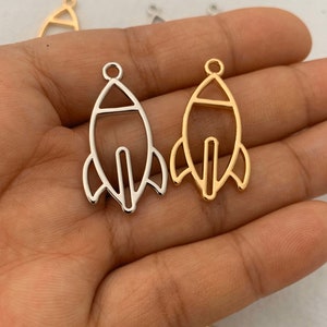 May include: Two metal rocket shaped charms, one gold and one silver. The charms are simple outlines of a rocket with a loop at the top for attaching to a necklace or bracelet.