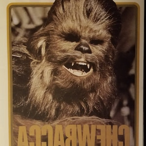 Könnte beinhalten: Eine Nahaufnahme von Chewbacca, einem Wookiee-Charakter aus Star Wars, mit einem braunen, pelzigen Gesicht und einem breiten, zahnigen Grinsen. Der Text "CHEWBACCA" ist in goldenen Buchstaben unter dem Bild geschrieben.
