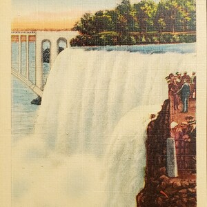 May include: A vintage postcard depicting the American Falls from Goat Island, Niagara Falls. The postcard features a view of the falls from the top of the island, with a bridge in the background. The falls are white and foamy, and the water is rushing over the edge of the cliff. The postcard is in good condition, with some minor wear and tear.