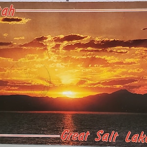 May include: A postcard featuring a sunset over the Great Salt Lake in Utah. The sky is a vibrant orange and yellow, with clouds reflecting the light. The mountains in the distance are silhouetted against the sky.