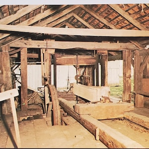 Può includere: Una segheria in legno vintage con una grande ruota in legno e una lunga tavola in legno. La segheria è all'interno di un edificio in legno con un tetto in legno.