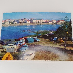 Pode incluir: Um cartão postal vintage mostrando uma vista panorâmica de uma cidade junto a um rio com vários carros e tendas montadas na margem do rio. O cartão postal está em bom estado com alguns sinais de desgaste.