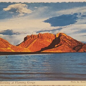 May include: A postcard featuring a scenic view of a red rock mountain range with a body of water in the foreground. The sky is blue with white clouds. The text on the postcard reads "Early Morning at Flaming Gorge" and "Photo by Stuart L. Tingley".