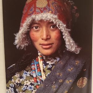 Puede incluir: Una mujer con un sombrero tradicional tibetano con ribete de piel blanca y una blusa con estampado floral de colores. Lleva un collar largo con cuentas de varios colores y un chal azul oscuro con estampado floral.