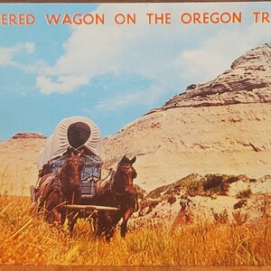 Könnte beinhalten: Ein Planwagen, gezogen von zwei Pferden, reist auf dem Oregon Trail. Der Wagen ist weiß mit einer Plane. Die Pferde sind braun und haben lange Mähnen. Im Hintergrund befindet sich eine große, felsige Klippe.